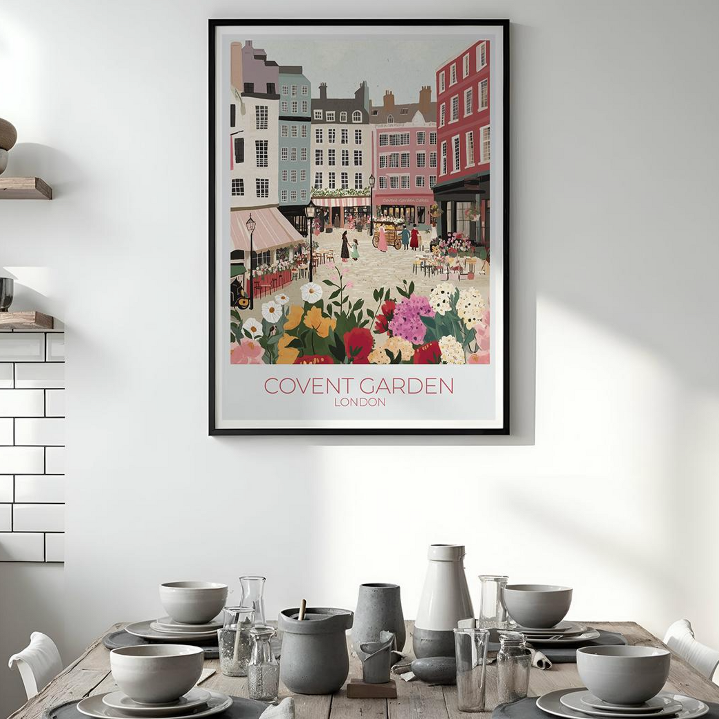 Framed illustration of Covent Garden, London, showing a vibrant market scene with colorful buildings, flower stalls, and people. The print hangs above a wooden dining table set with kitchenware, with white subway tiles and shelves in the background. Text on the print reads 'COVENT GARDEN LONDON'.