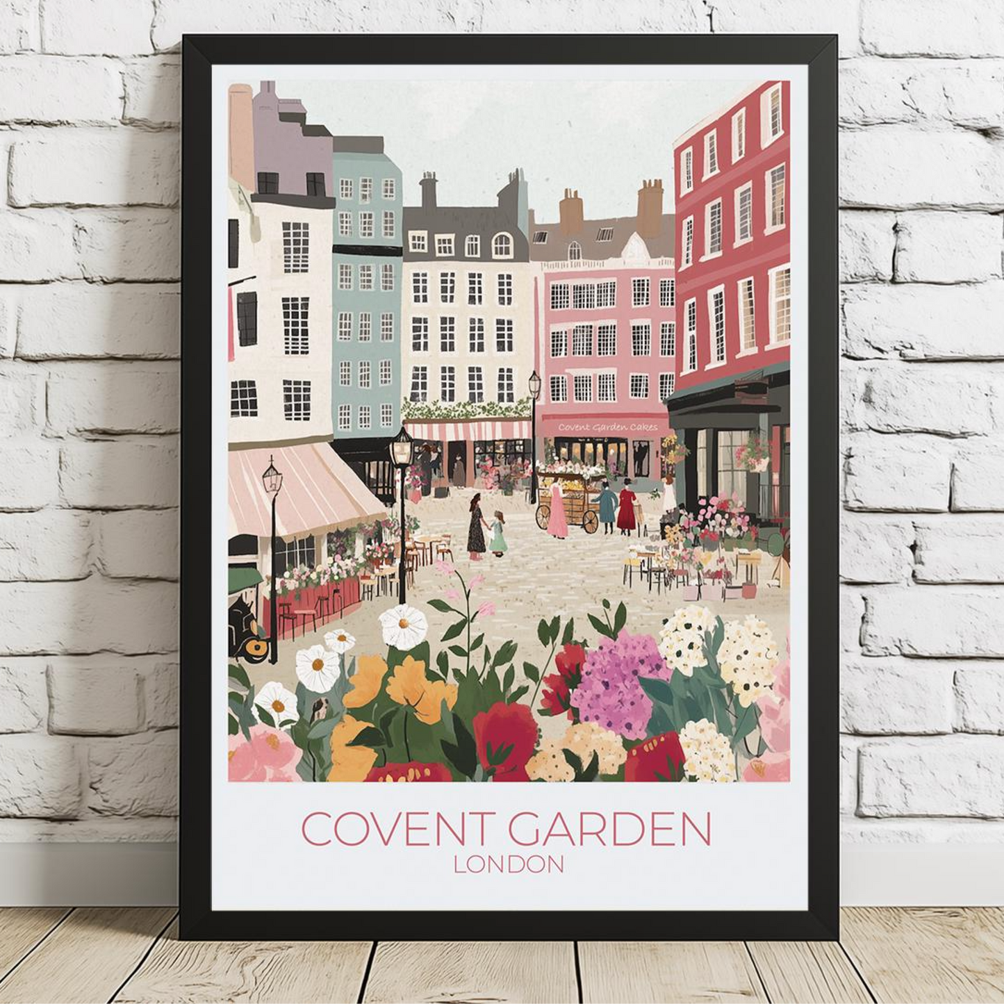 Framed illustration of Covent Garden, London, showing a vibrant market scene with colorful buildings, flower stalls, and people. The print hangs above a wooden dining table set with kitchenware, with white subway tiles and shelves in the background. Text on the print reads 'COVENT GARDEN LONDON'.