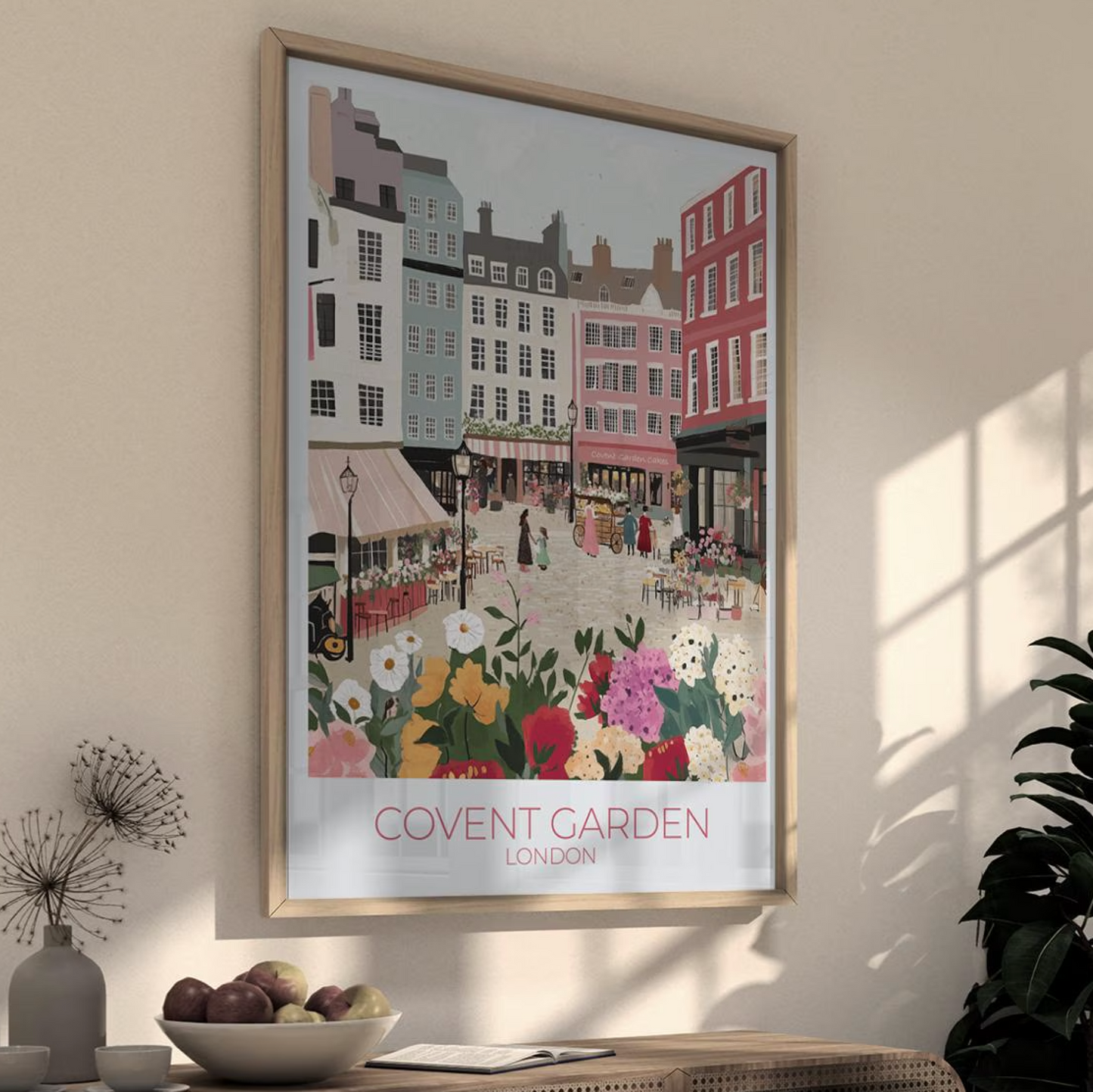 Framed illustration of Covent Garden, London, showing a vibrant market scene with colorful buildings, flower stalls, and people. The print hangs above a wooden dining table set with kitchenware, with white subway tiles and shelves in the background. Text on the print reads 'COVENT GARDEN LONDON'.