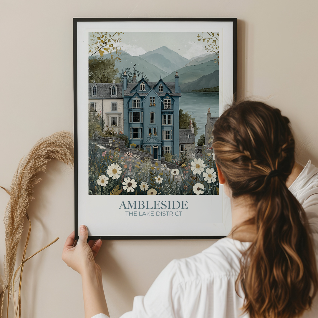 Woman holding a framed print of a scenic house with mountains and flowers, titled 'Ambleside The Lake District'.